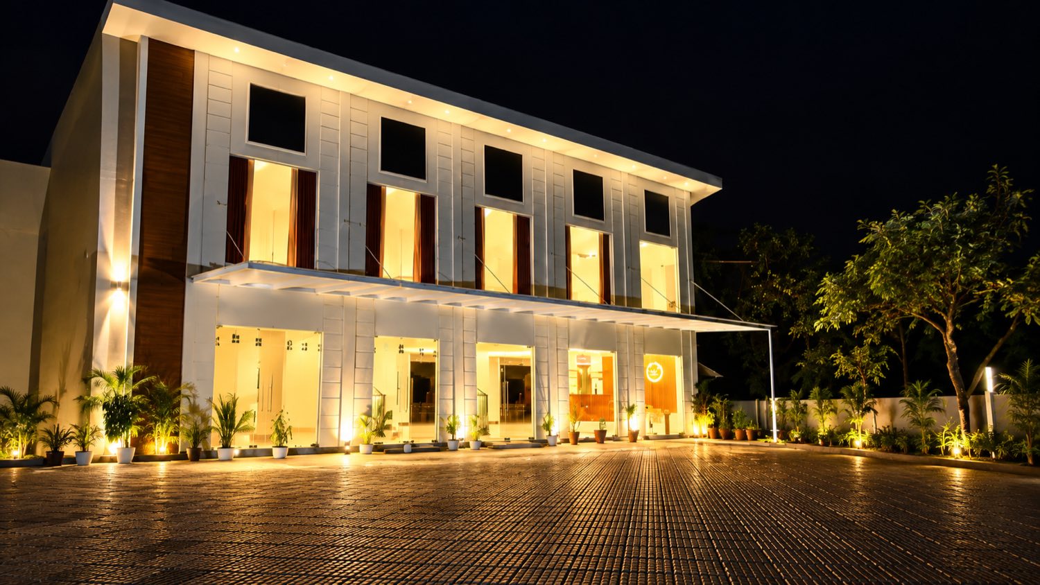 The Vanyora hotel building lit at night: a white two-storey block with a deep verandah, warm light in every window, potted palms along a paved forecourt.