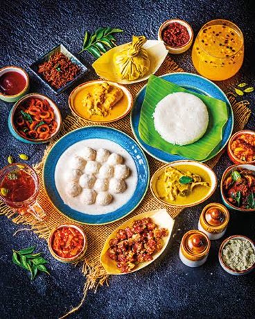A spread of Kerala nadan dishes laid out together: kappa, fish curry, thoran, appam, pickles and black tea.